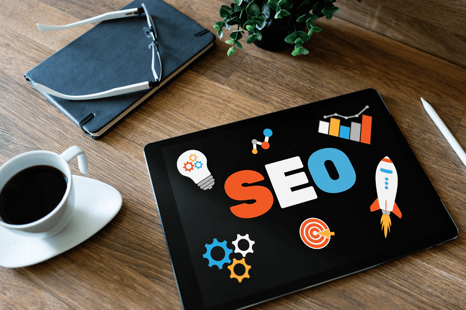Picture of office desk with coffee, notebook, and tablet that displays the word "SEO". Picture of office desk with coffee, notebook, and tablet that displays the word "SEO".